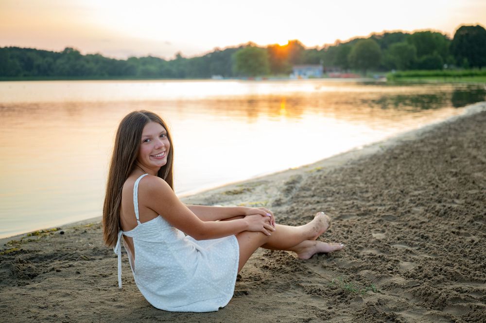 Young girl in white dress sitting barefoot on sandy beach by calm lake at golden sunset.