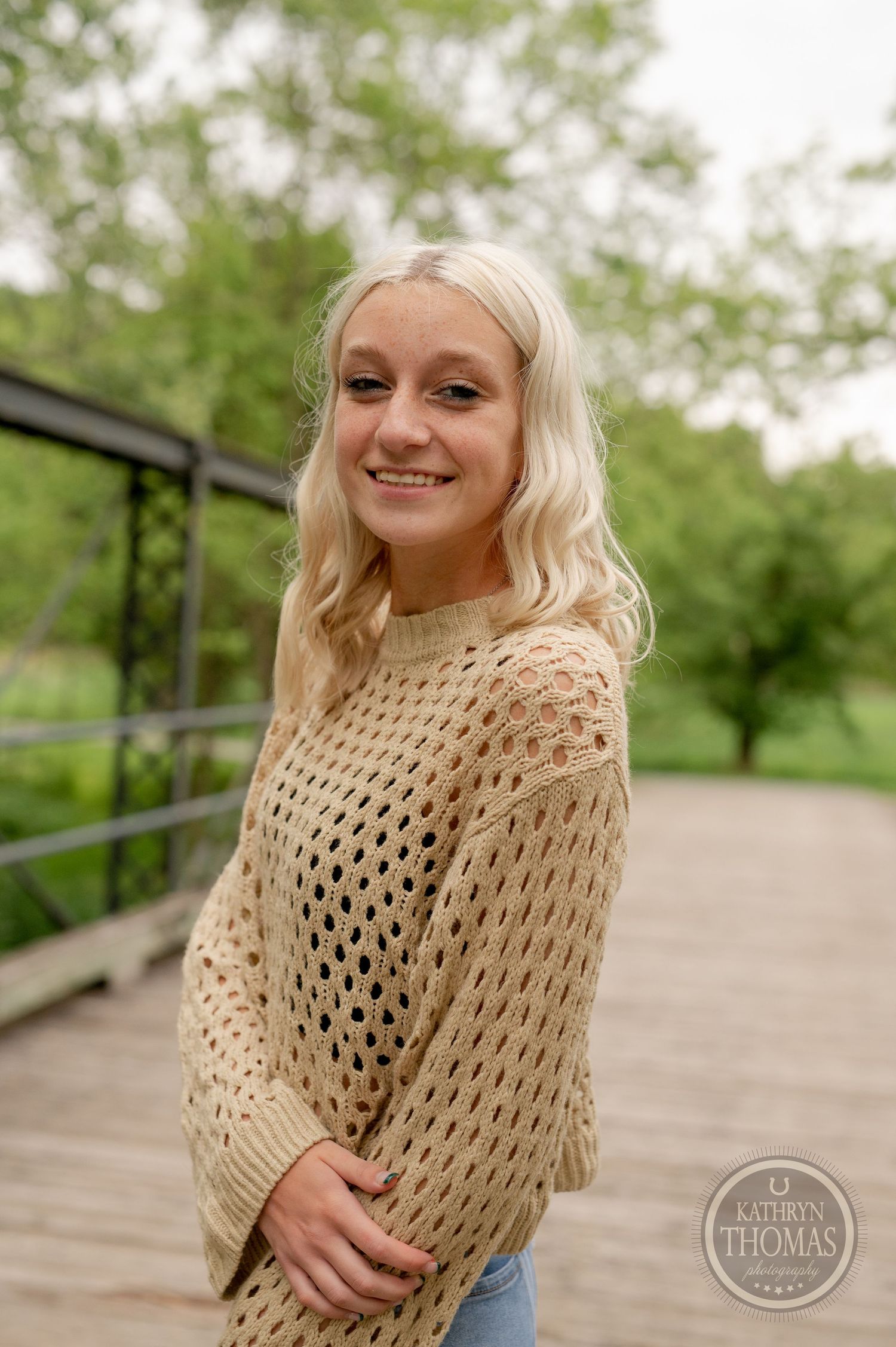 Lifestyle portrait showing open-knit cream sweater against lush green background on wooden bridge.