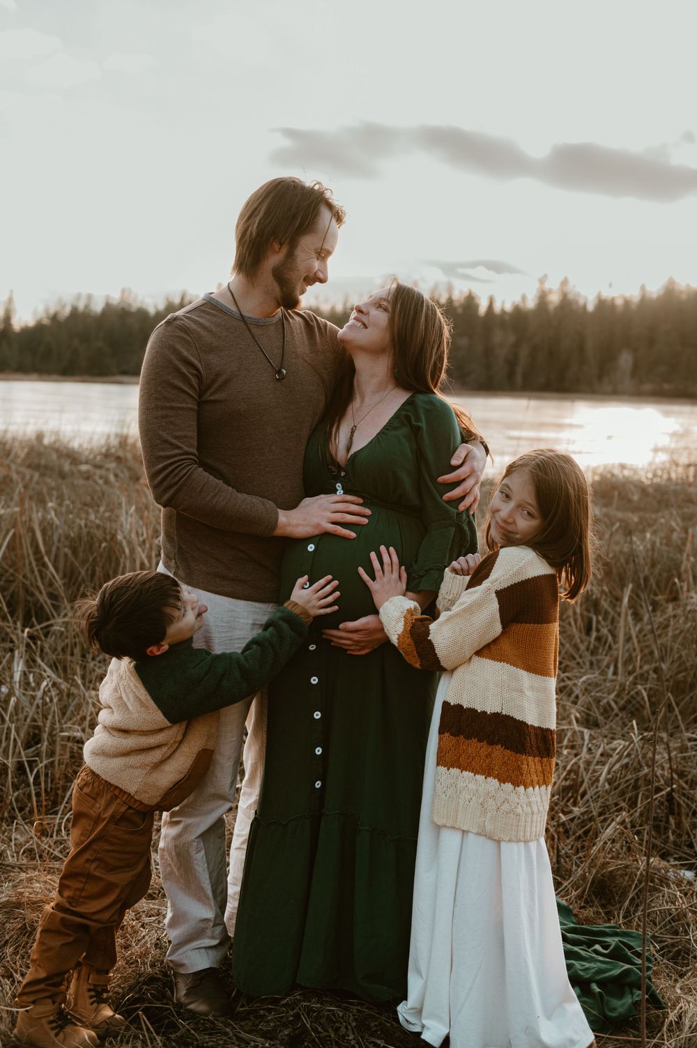 A family shares a tender moment together at sunset by a lake, with the mother wearing a long green dress.