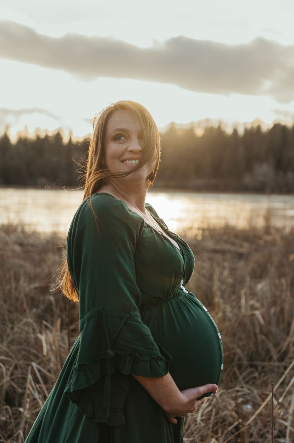 Beautiful maternity photo in emerald green dress with glowing sunset light over lake and tall grasses in background.