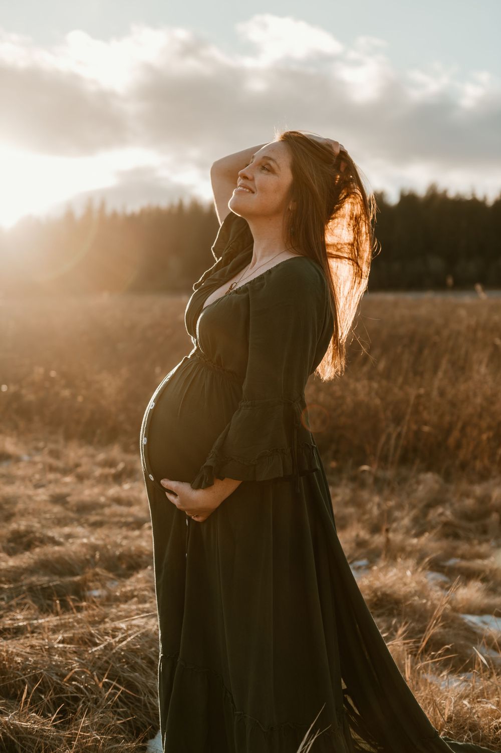 Pregnant woman in black dress stands in golden sunlit field at sunset with flowing hair in the breeze.