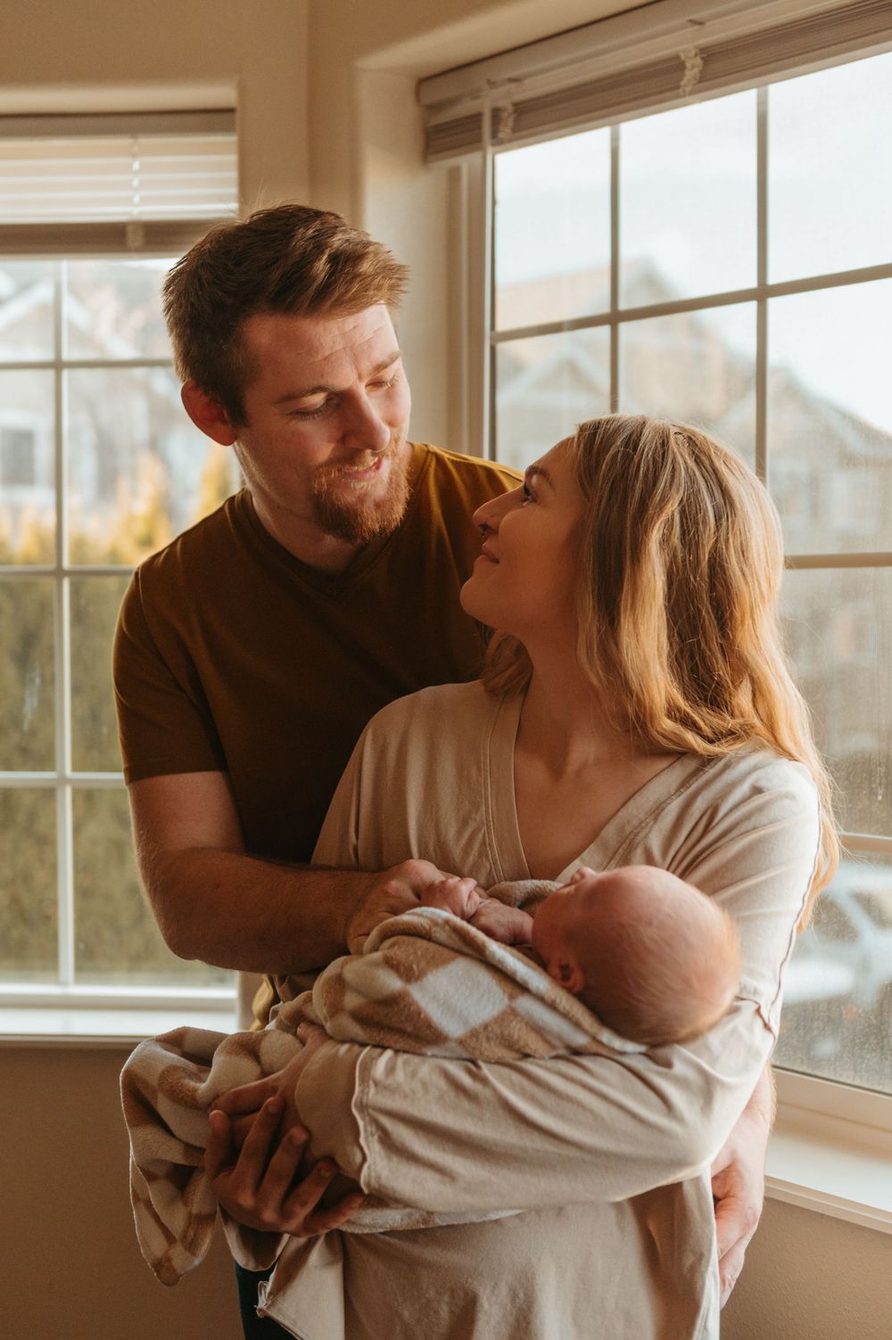 A couple lovingly holds their newborn baby while standing by a bright window in a warm-lit room.