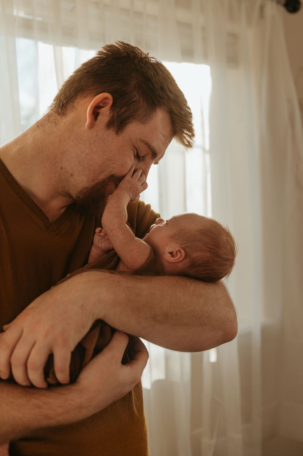 A tender moment between a parent and newborn baby in warm lighting by a window with sheer curtains.