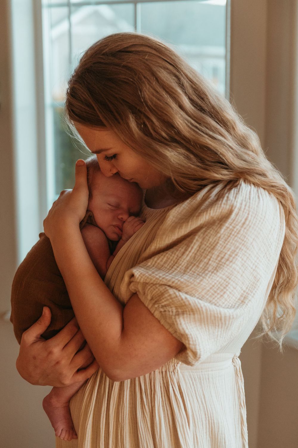 A tender moment between mother and newborn captured in soft natural light by a window.
