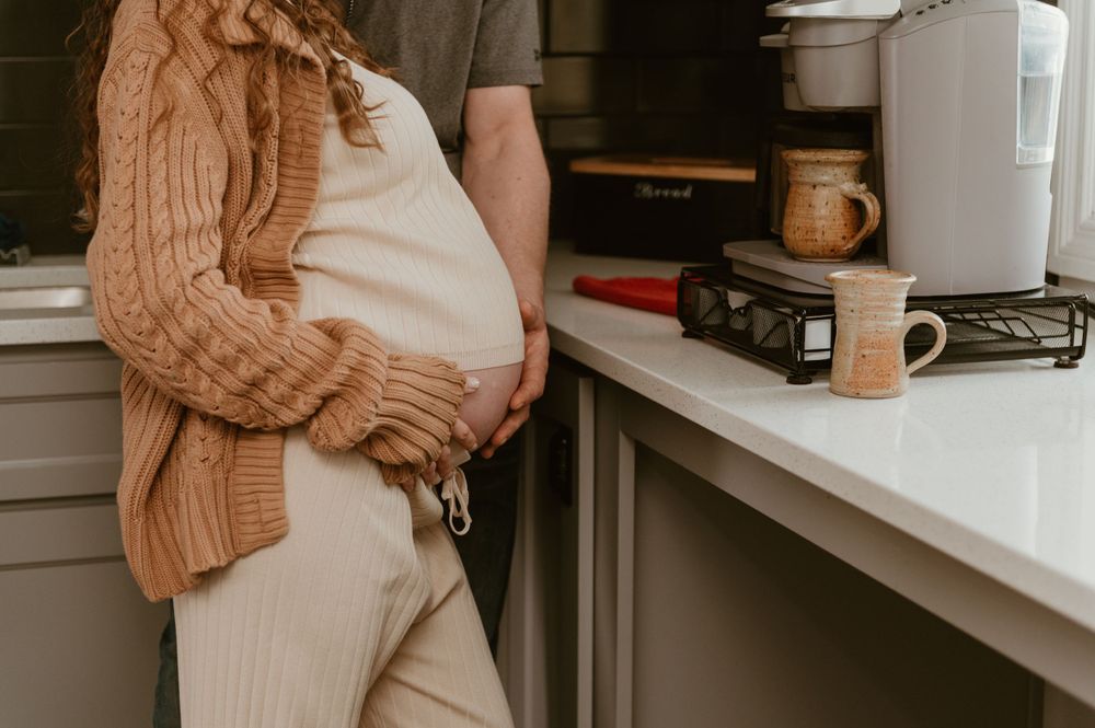 Someone wearing a cozy beige sweater cradles their pregnant belly in a sunlit kitchen setting.