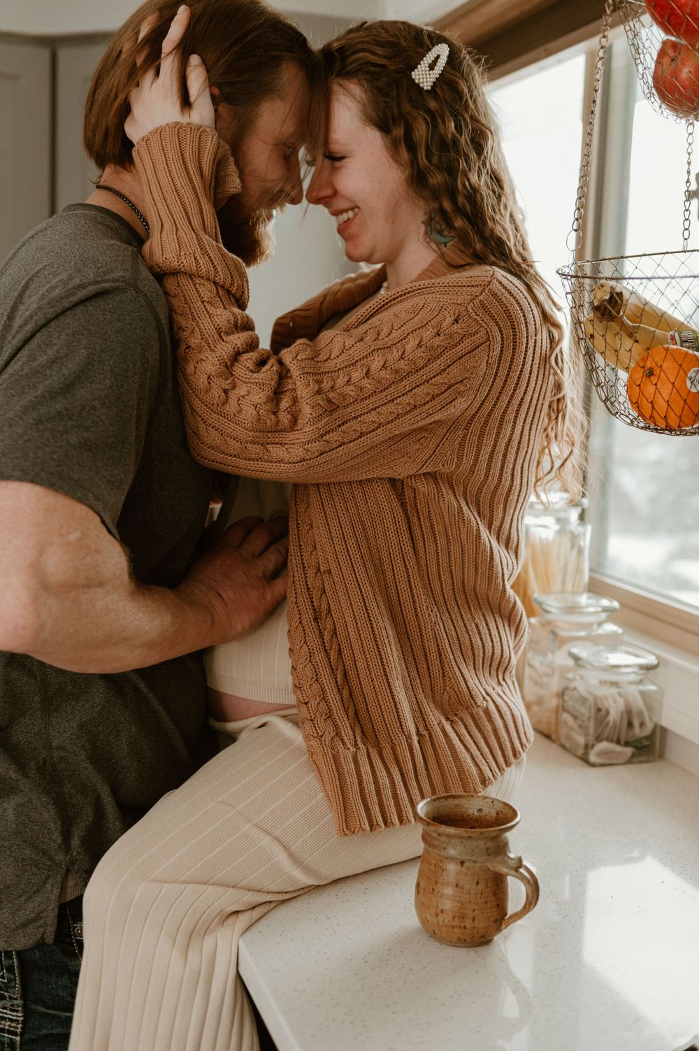 Couple in cozy sweaters share an intimate moment in sunlit kitchen with ceramic mug on counter.