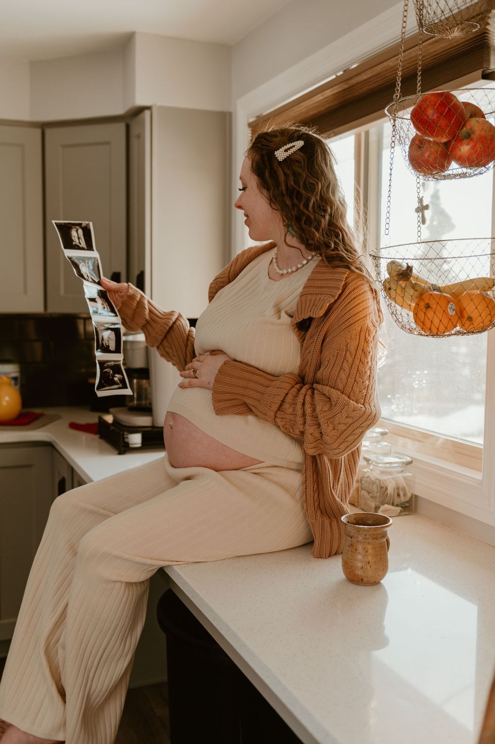 A pregnant woman in white sits on a counter looking at photos while wearing a cozy brown cardigan in a sunlit kitchen.