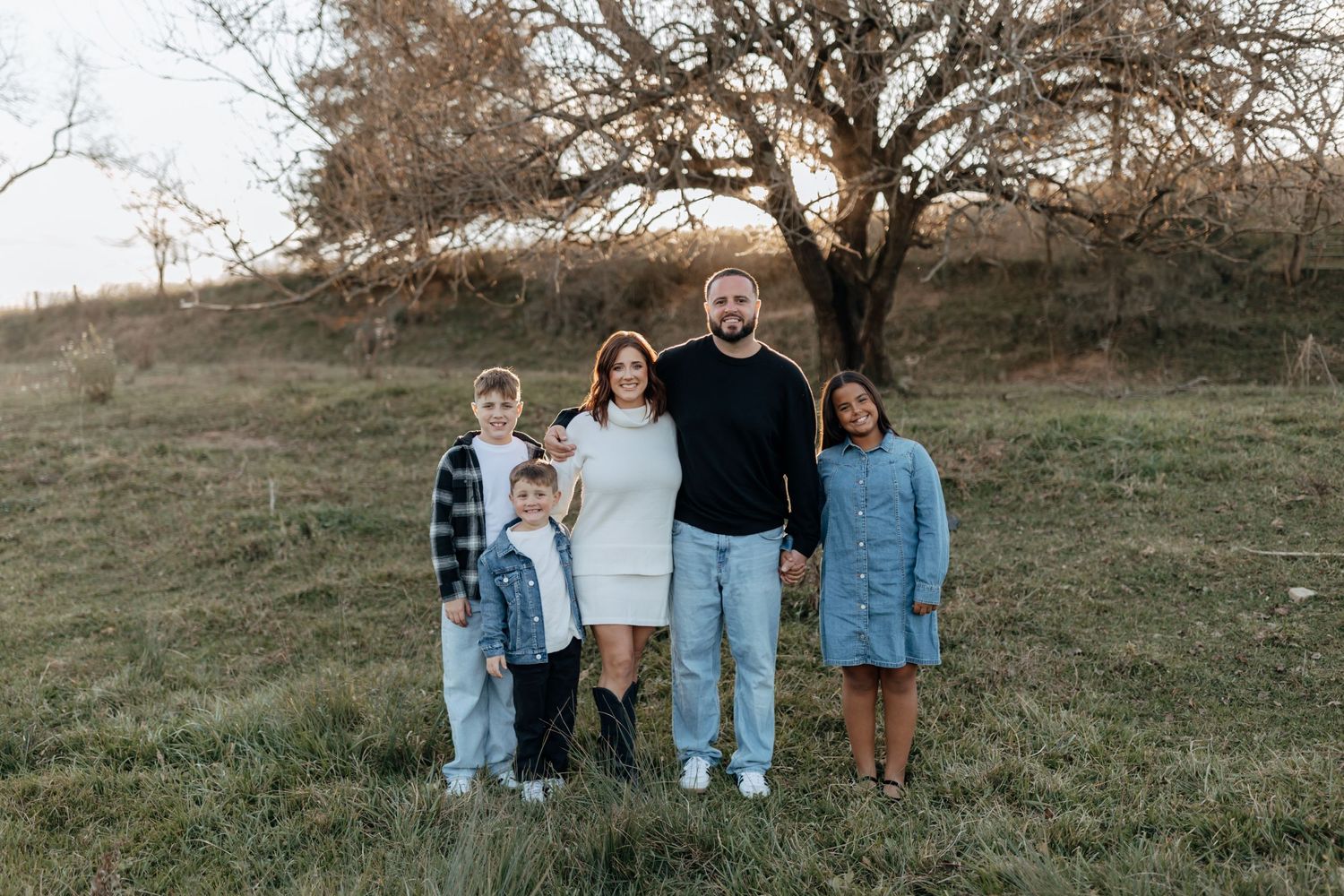 A family poses together for a casual outdoor portrait against a rustic backdrop with a bare tree.