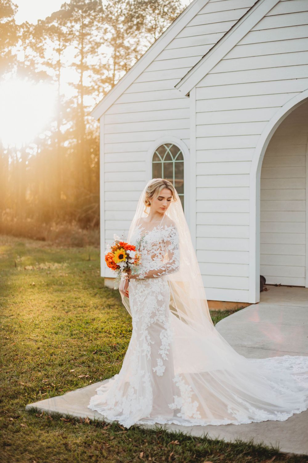 A wedding dress photo featuring delicate lace sleeves and vibrant orange bouquet against white chapel backdrop.