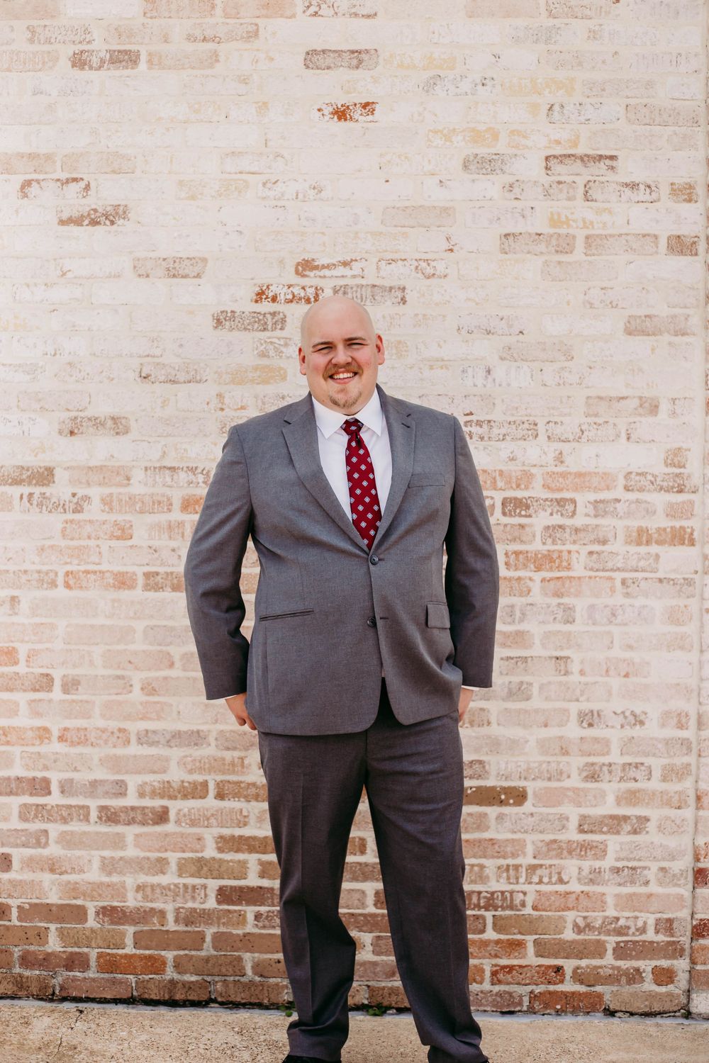 Professional portrait of man in grey suit and red tie against textured brick wall.