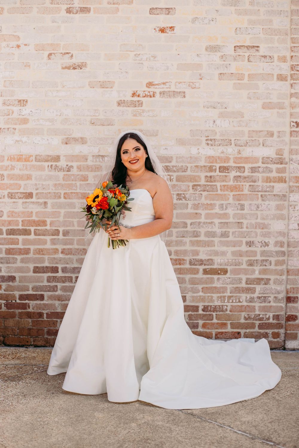 Classic bridal portrait with strapless ballgown and sunflower bouquet against brick wall backdrop.