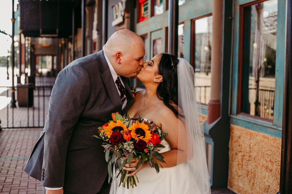 Newlyweds kiss while holding vibrant sunflower bouquet under storefront awning.