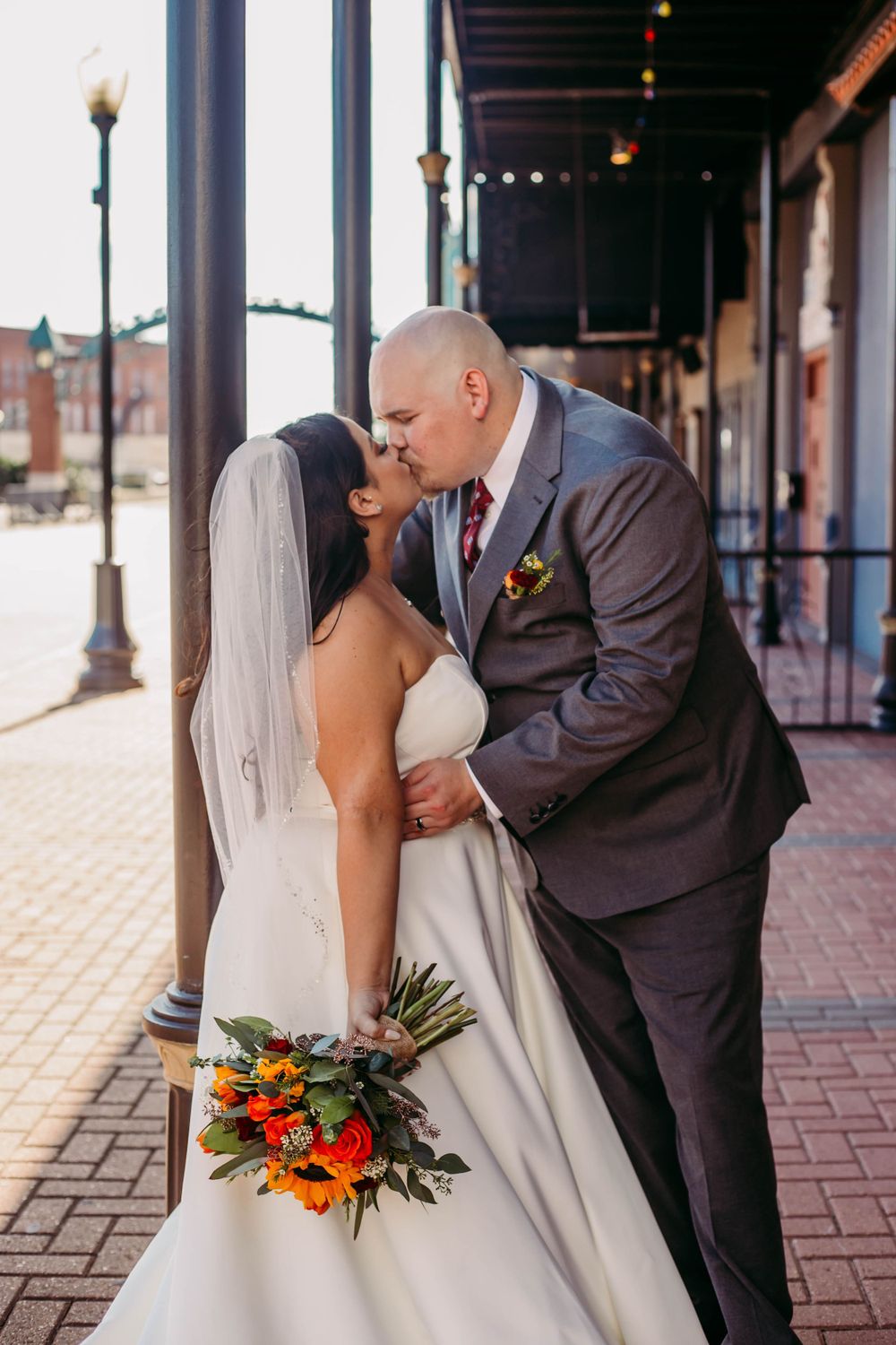 Couple sharing a romantic kiss under a covered walkway with brick pavement and decorative street lamps.