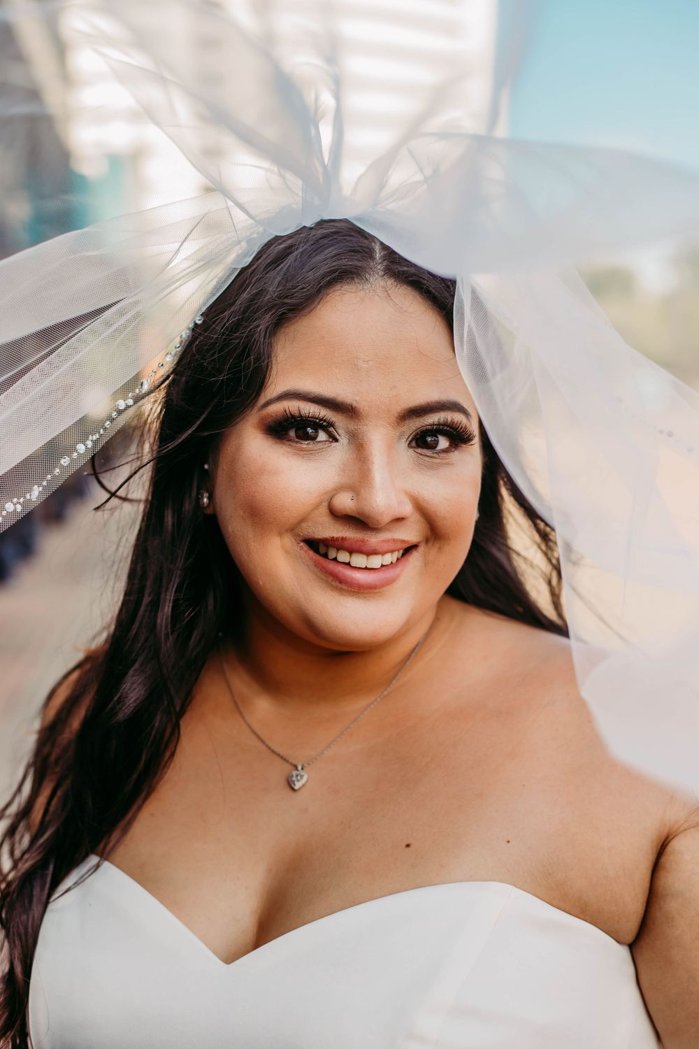 Bride smiling with flowing veil against blue sky on wedding day.