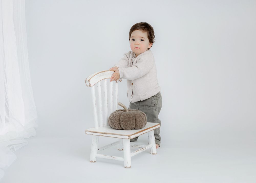 A toddler in gray pants and cream sweater plays with a white chair.