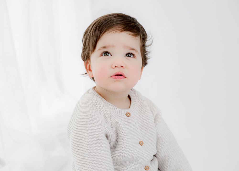 A young child wearing a cream cardigan poses in a bright studio setting.