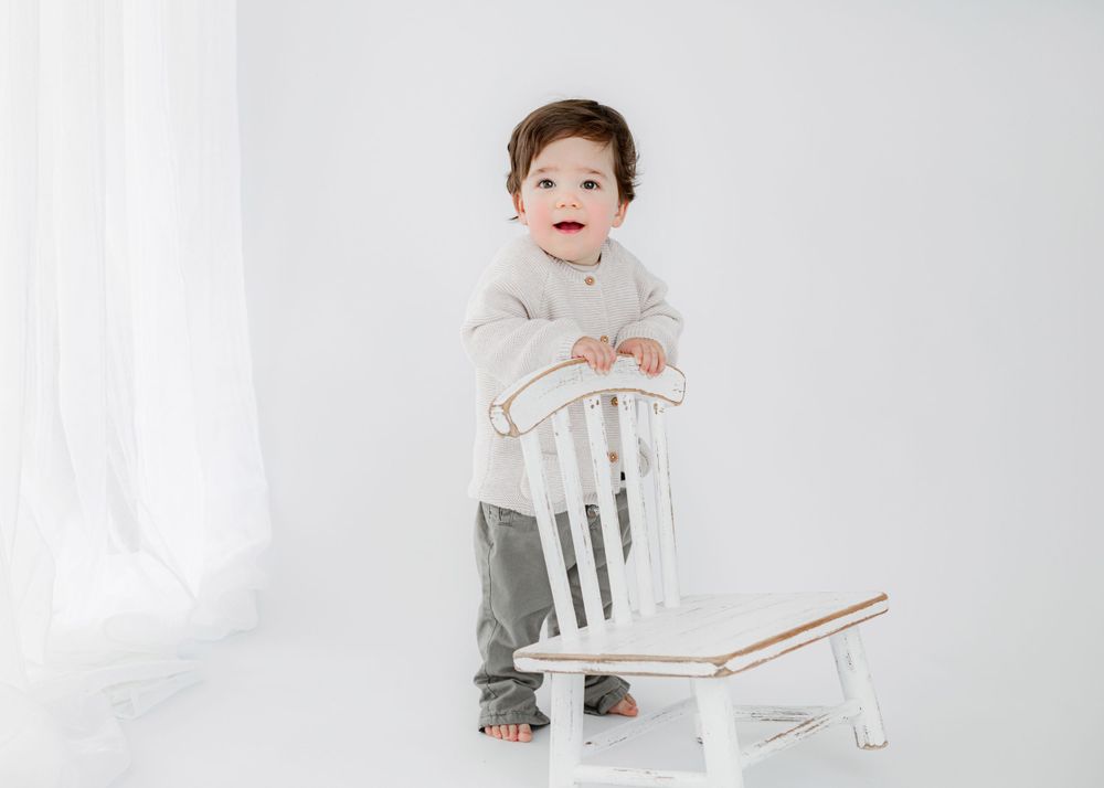 A small child stands beside a white wooden chair in a minimalist setting.