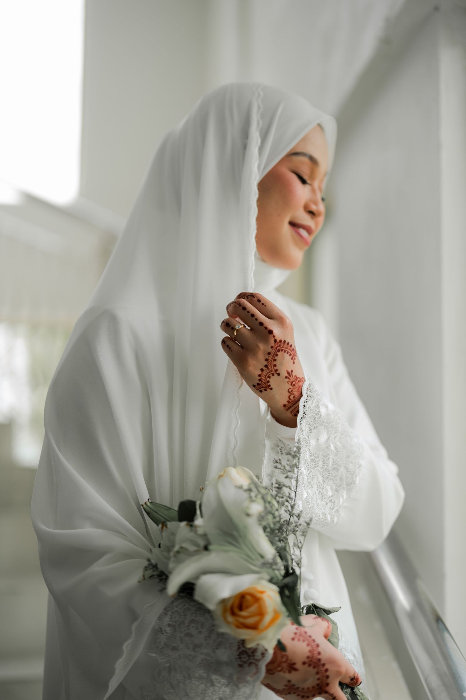 Side profile of bride in white hijab holding flowers, showing intricate henna designs on hands.