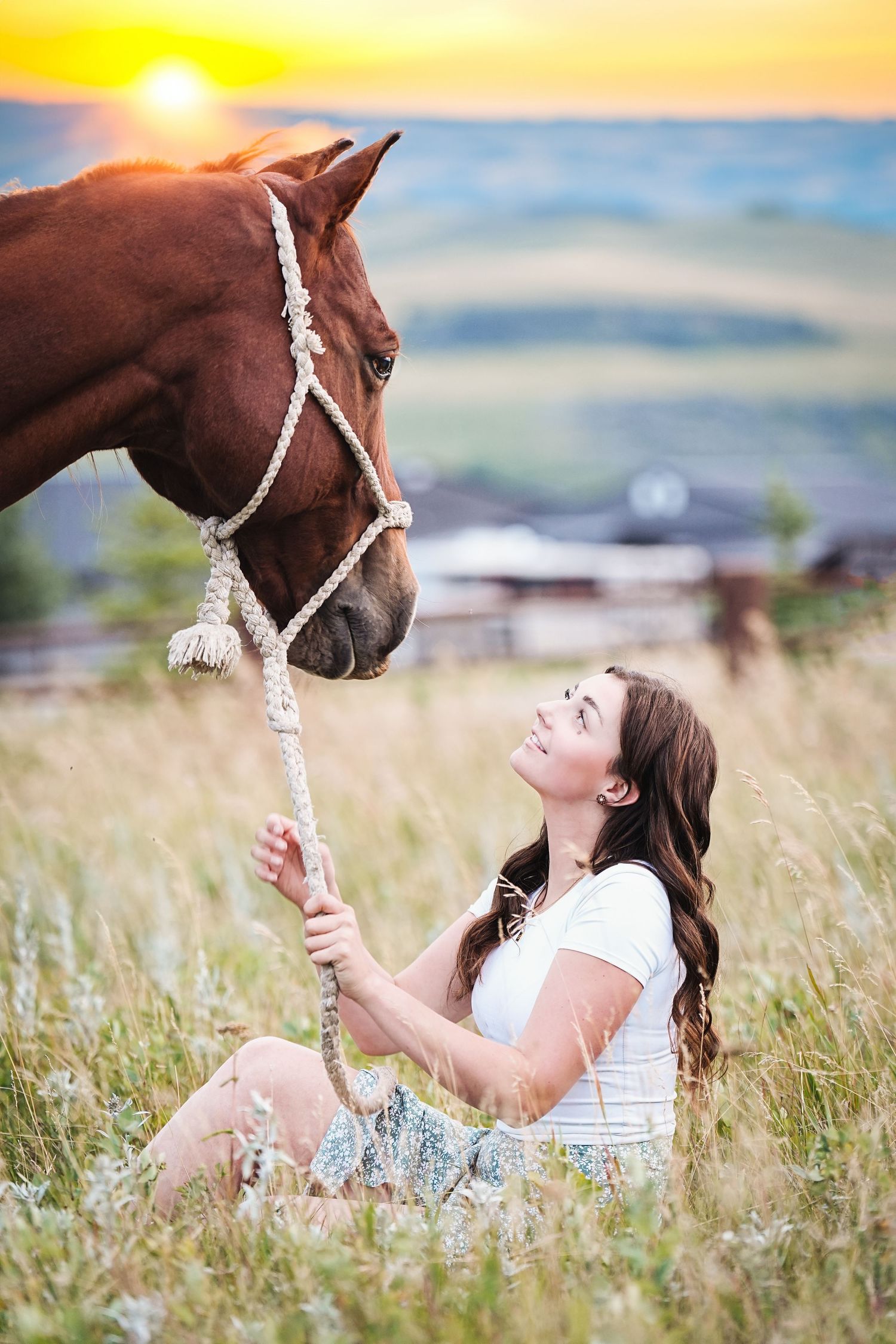 Young woman sitting in meadow connects with brown horse against sunset sky.