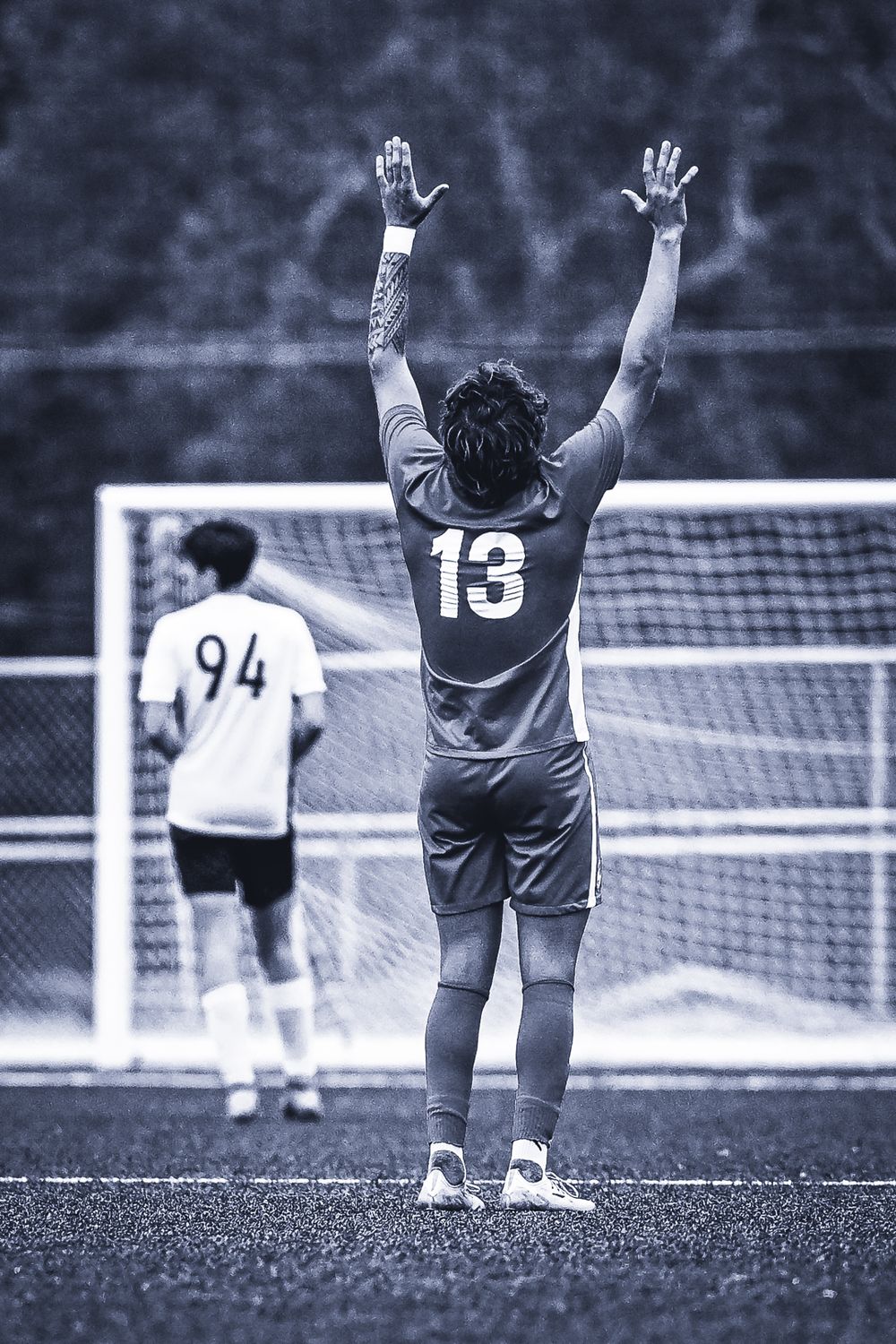 A soccer goalkeeper wearing jersey number 13 stands with raised arms on a sports field in black and white.