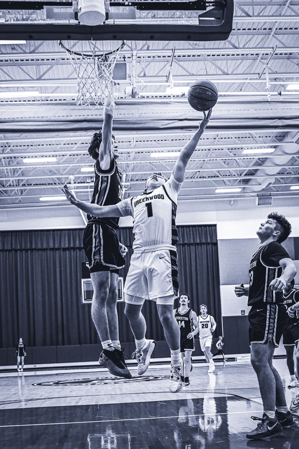 Indoor basketball players leap for a shot during an intense game on a glossy court underneath metal rafters.