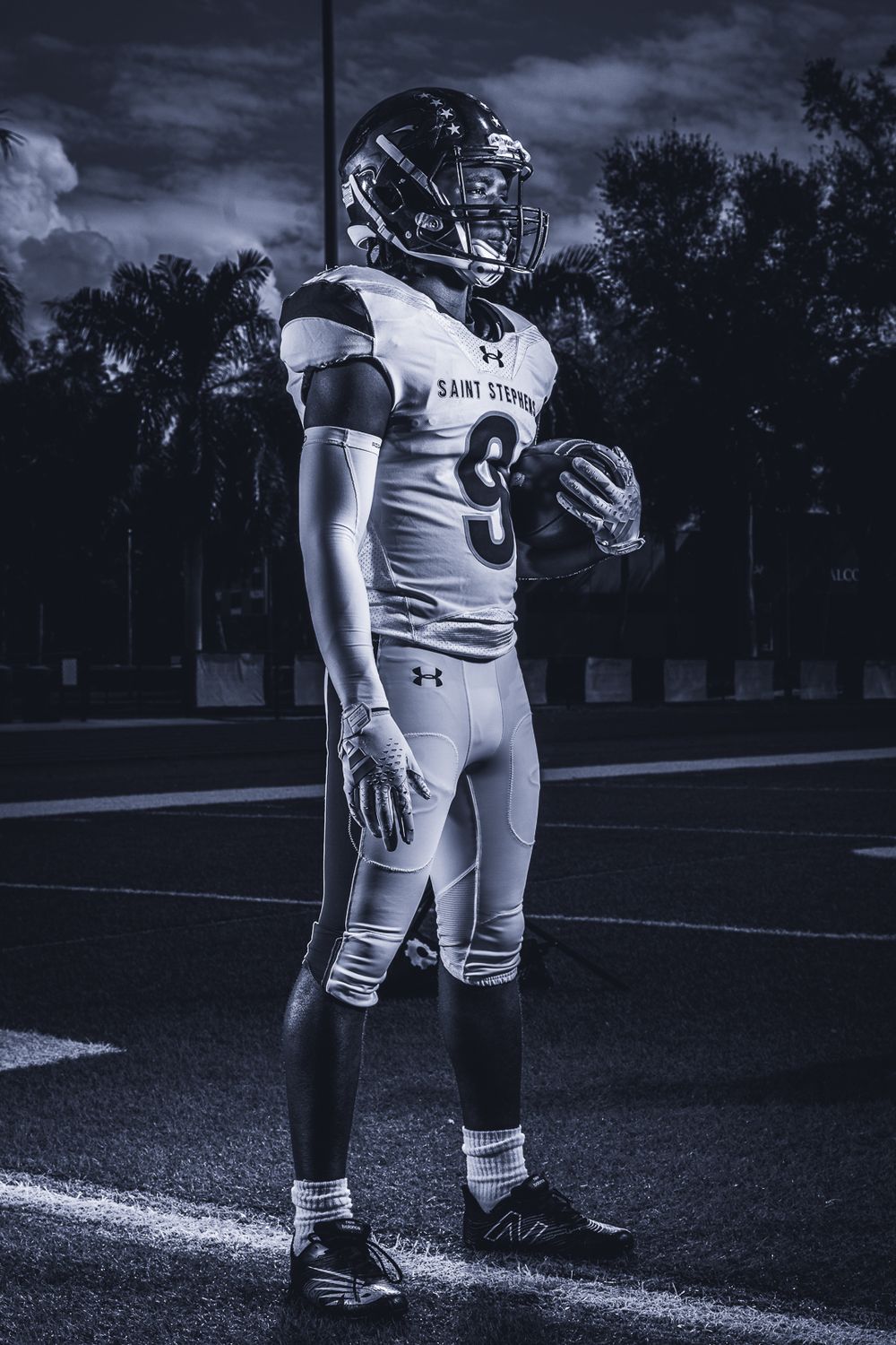 Football player in white Under Armour uniform stands on field at dusk in dramatic black and white photo.