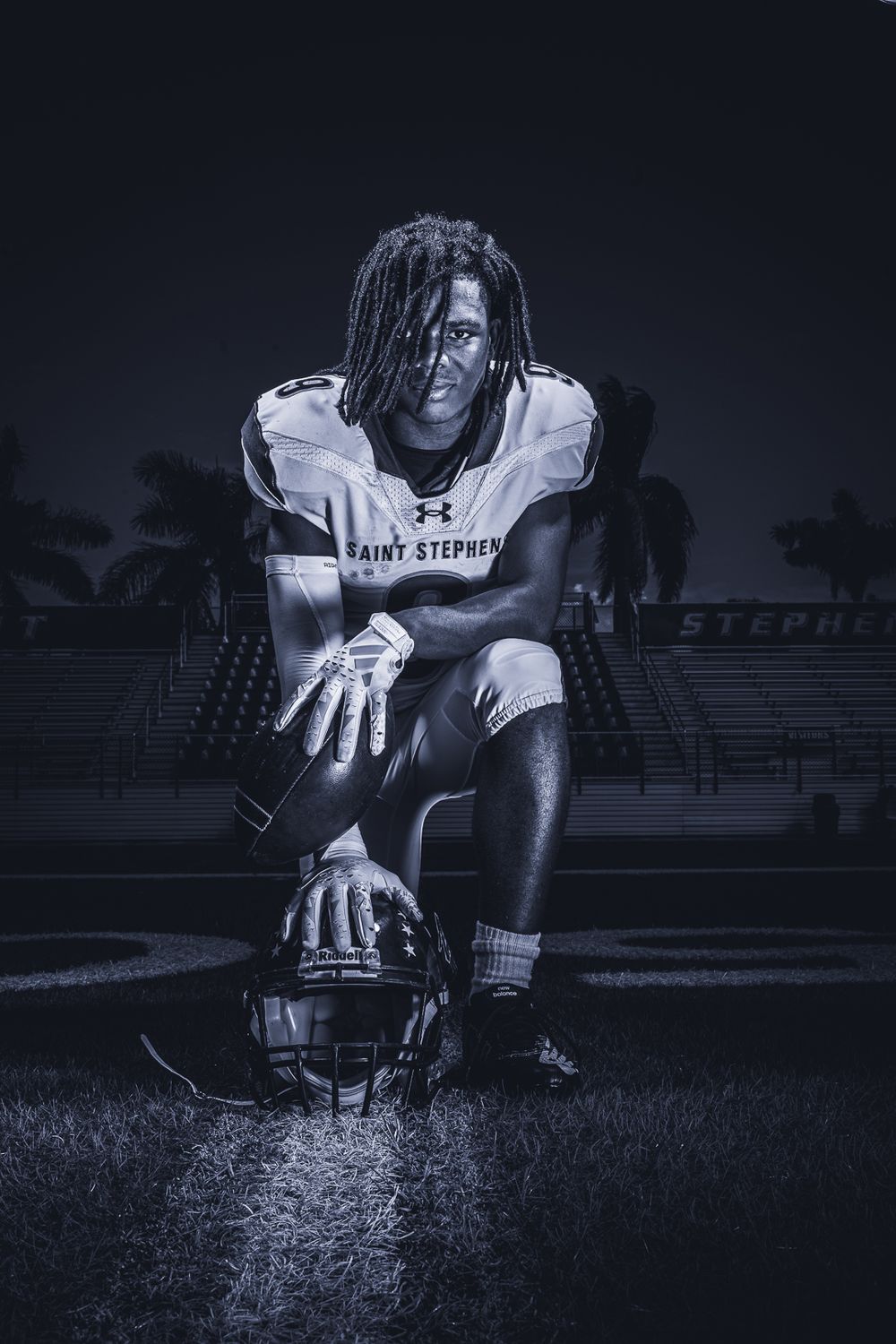 Football player in white uniform kneels holding helmet in dramatic nighttime portrait.