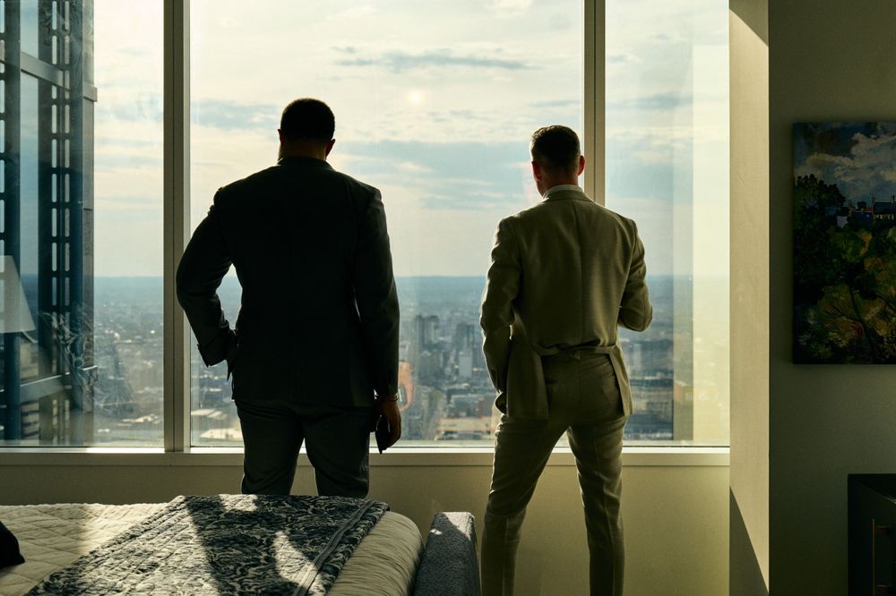 Two silhouettes stand by large windows overlooking a cityscape at sunset.