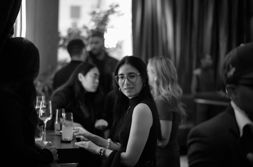 A black and white photo of patrons dining at a dimly lit restaurant bar with wine glasses on the table.