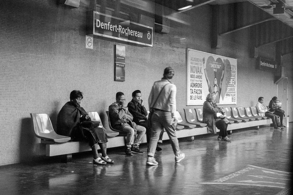 Passengers waiting in subway station with posters on tiled wall.