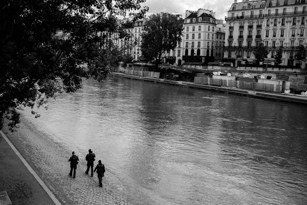 Two figures walking along Seine River with Parisian buildings in background.