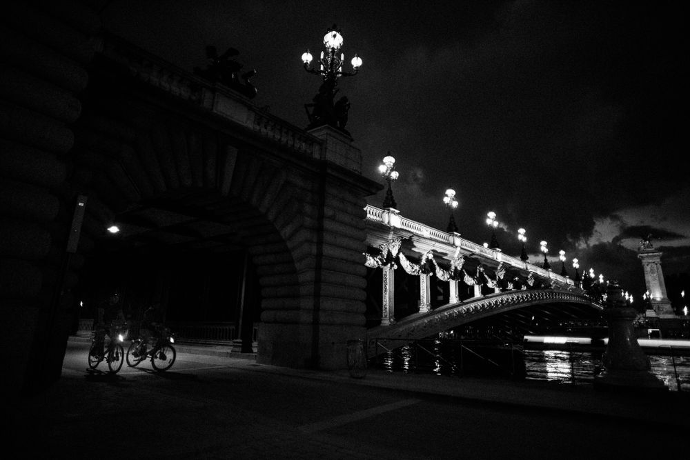 Illuminated bridge with ornate lampposts and railings reflected in river water at night in black and white.
