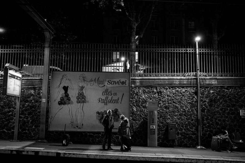 People standing by illuminated street lamp near graffitied wall at night.