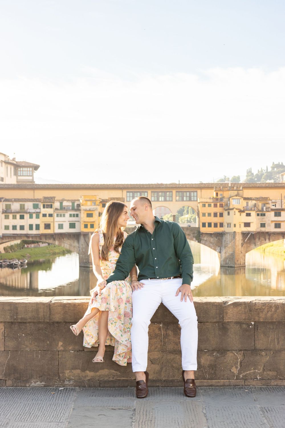 A couple sits on a stone wall overlooking the iconic Ponte Vecchio bridge in Florence.