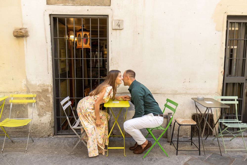 A couple shares an intimate moment at a small yellow cafe table with green chairs on a European street.