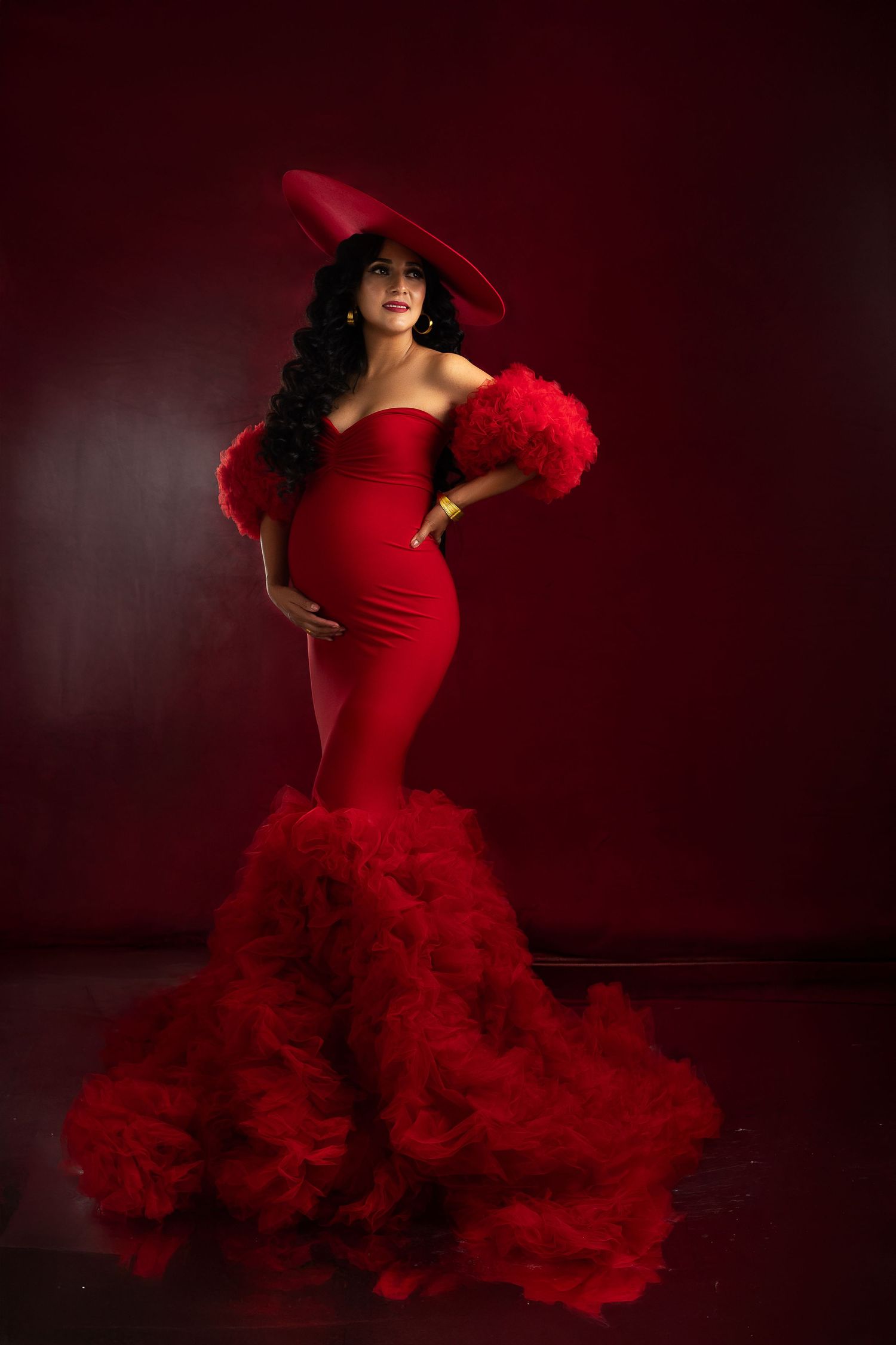 Dramatic maternity photo featuring a red gown with flowing tulle and matching wide-brim hat.