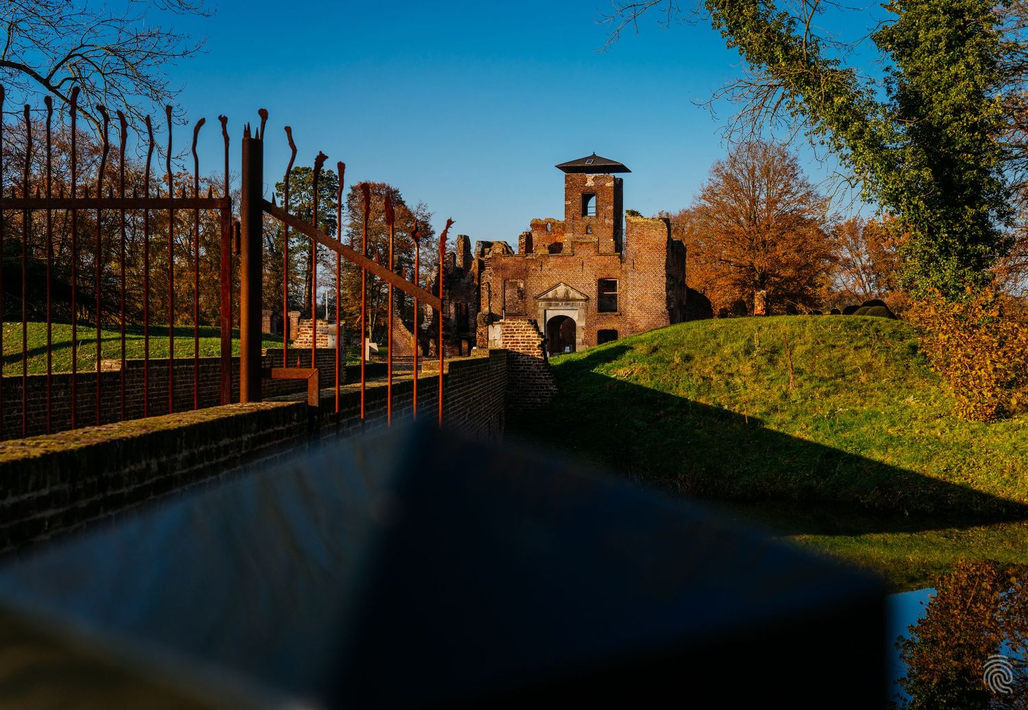 Wide-angle photo of a path leading through iron gates towards a stone castle or historic building. Kasteel Bleijenbeek