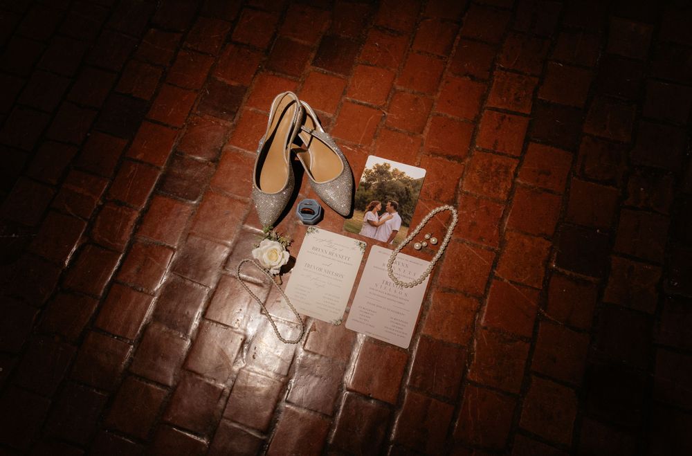 Bridal accessories including heels, jewelry, and stationery arranged on rustic brick floor.