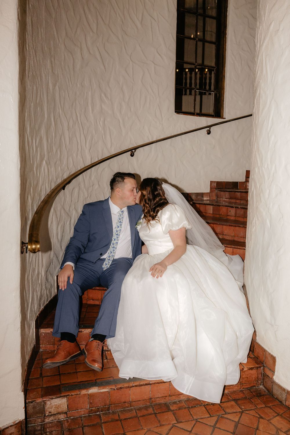 Bride in white gown and groom in blue suit kissing on terracotta spiral staircase.