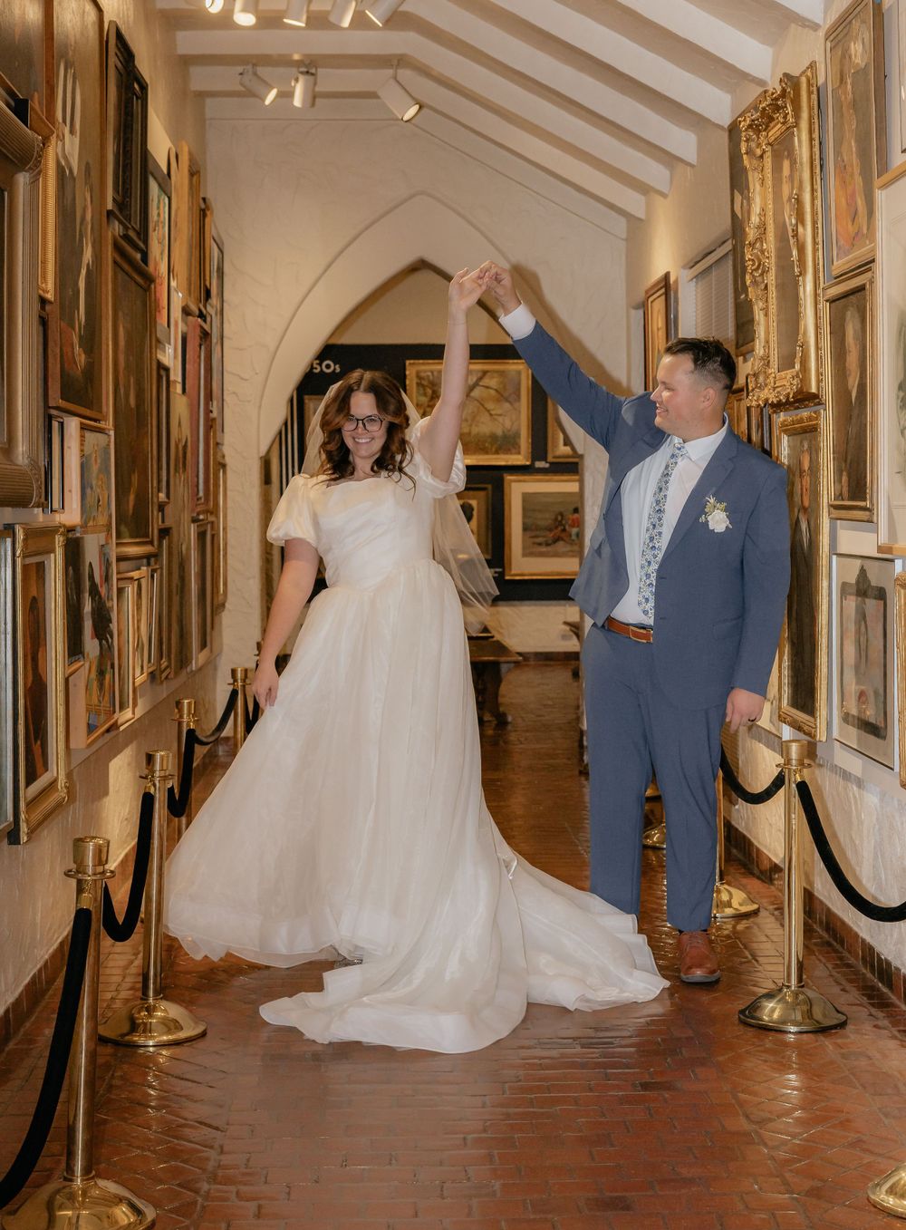 Bride and groom dancing joyfully through an art gallery corridor lined with framed paintings.