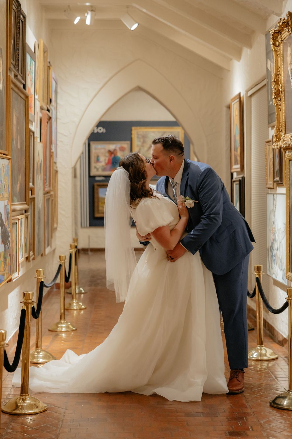 Bride and groom sharing a romantic kiss in an elegant art gallery hallway with paintings.