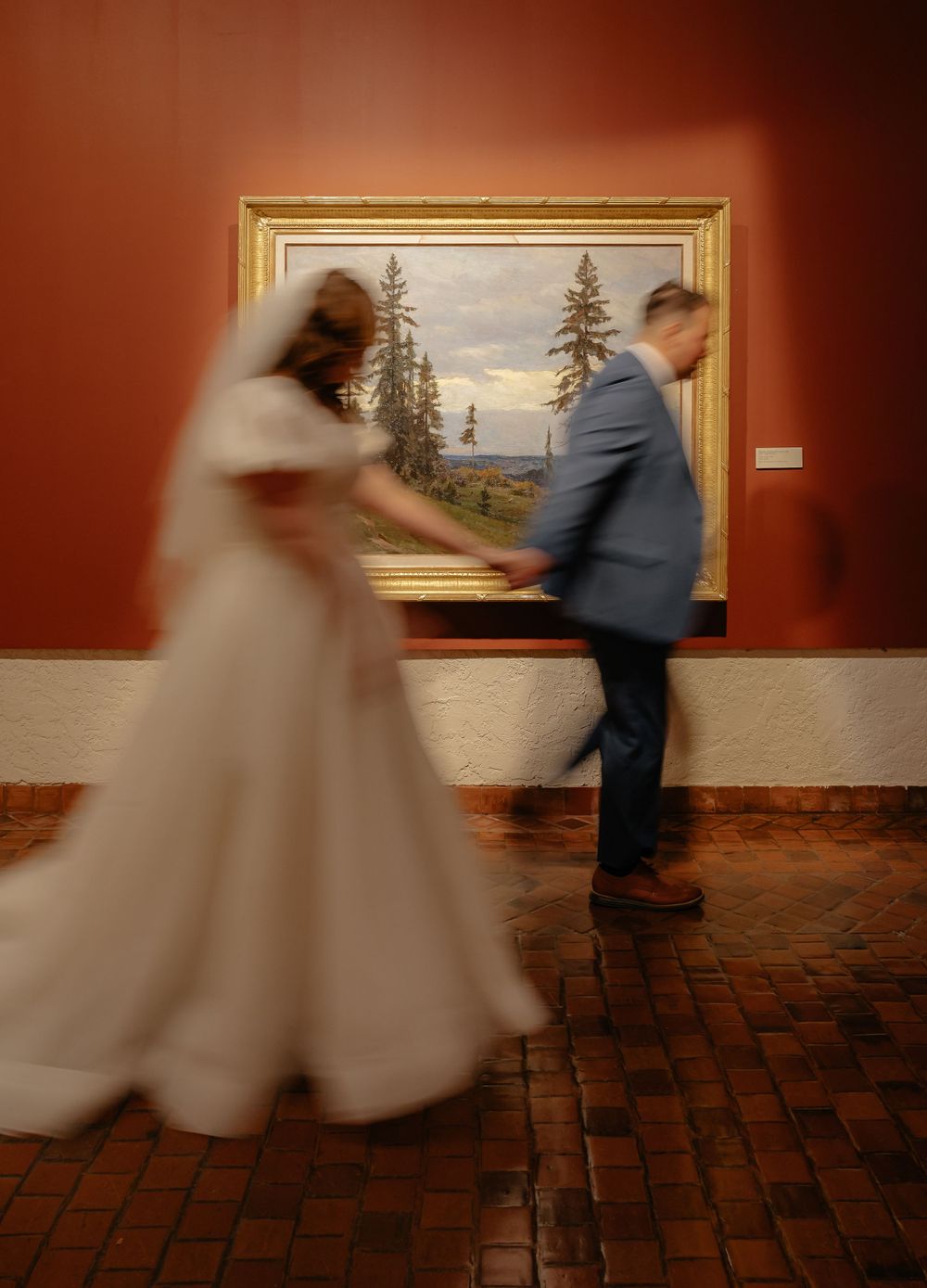 Bride and groom walk past a landscape painting in a red-walled art gallery wedding venue.