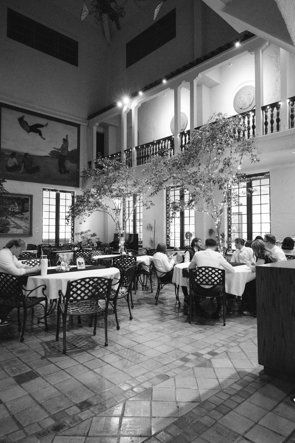 Black and white photo of wedding reception guests dining in a grand historic venue with balcony.
