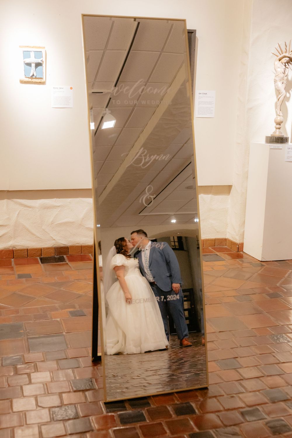 Bride and groom share a kiss reflected in a decorative gold mirror engraved with wedding details.