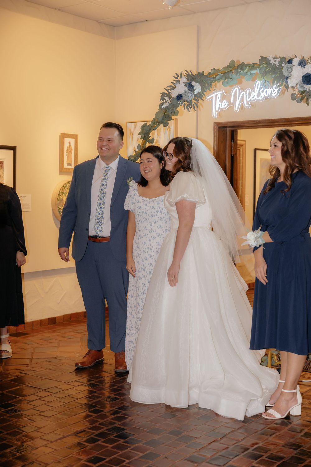 Smiling bride and groom walk through art gallery reception hall beneath The Nielsons neon sign.