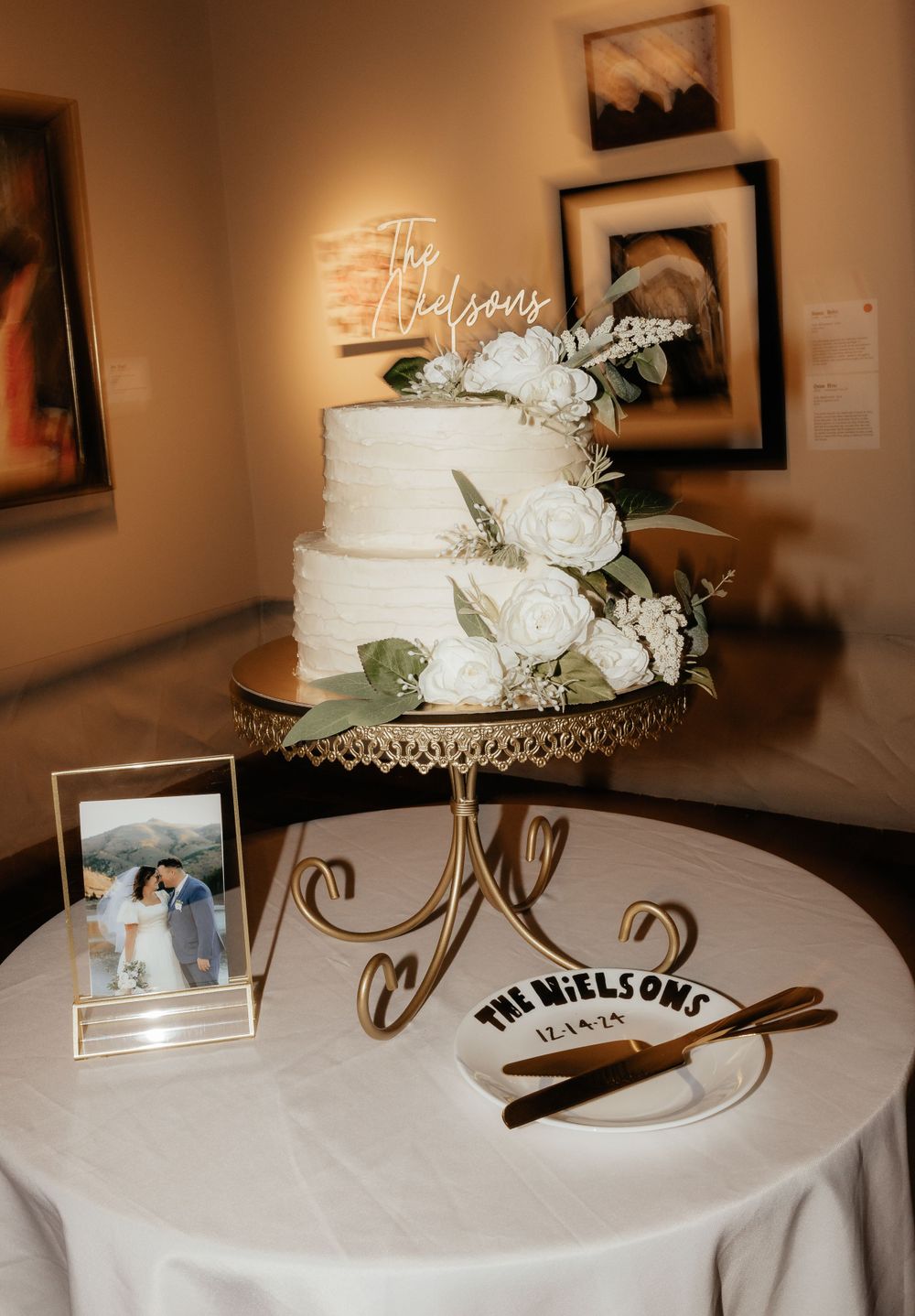 Two-tier white wedding cake with roses on gold stand beside The Nielsons personalized cake board.