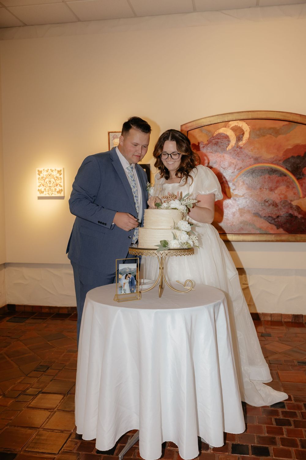 Bride and groom cut their white floral wedding cake together in front of a colorful mural painting.