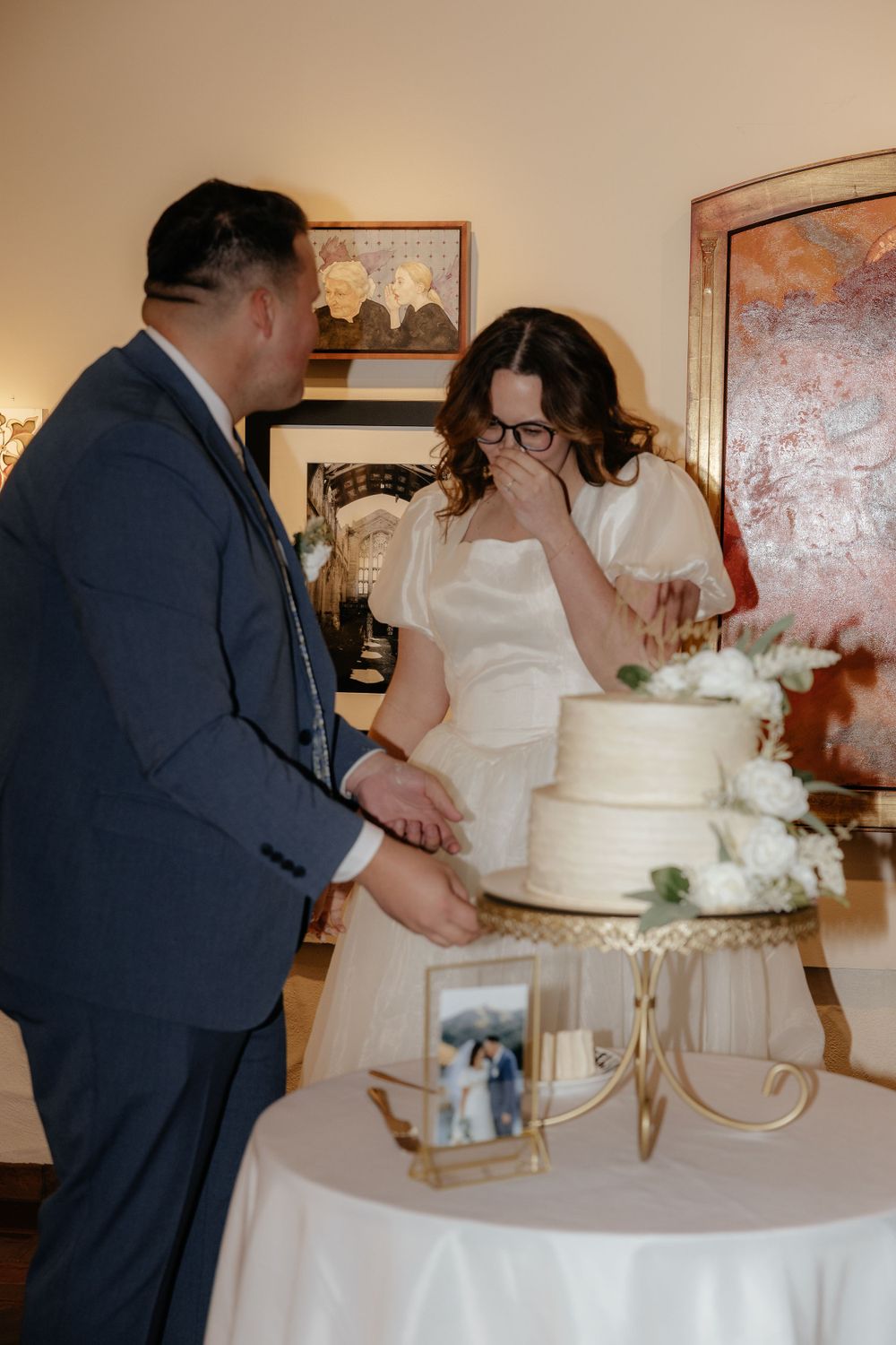 Bride laughing while cutting wedding cake with groom at elegant art gallery reception.