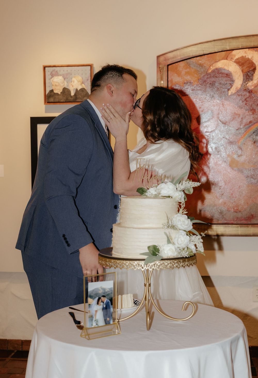Newlyweds sharing a kiss behind their elegant white wedding cake at their reception.