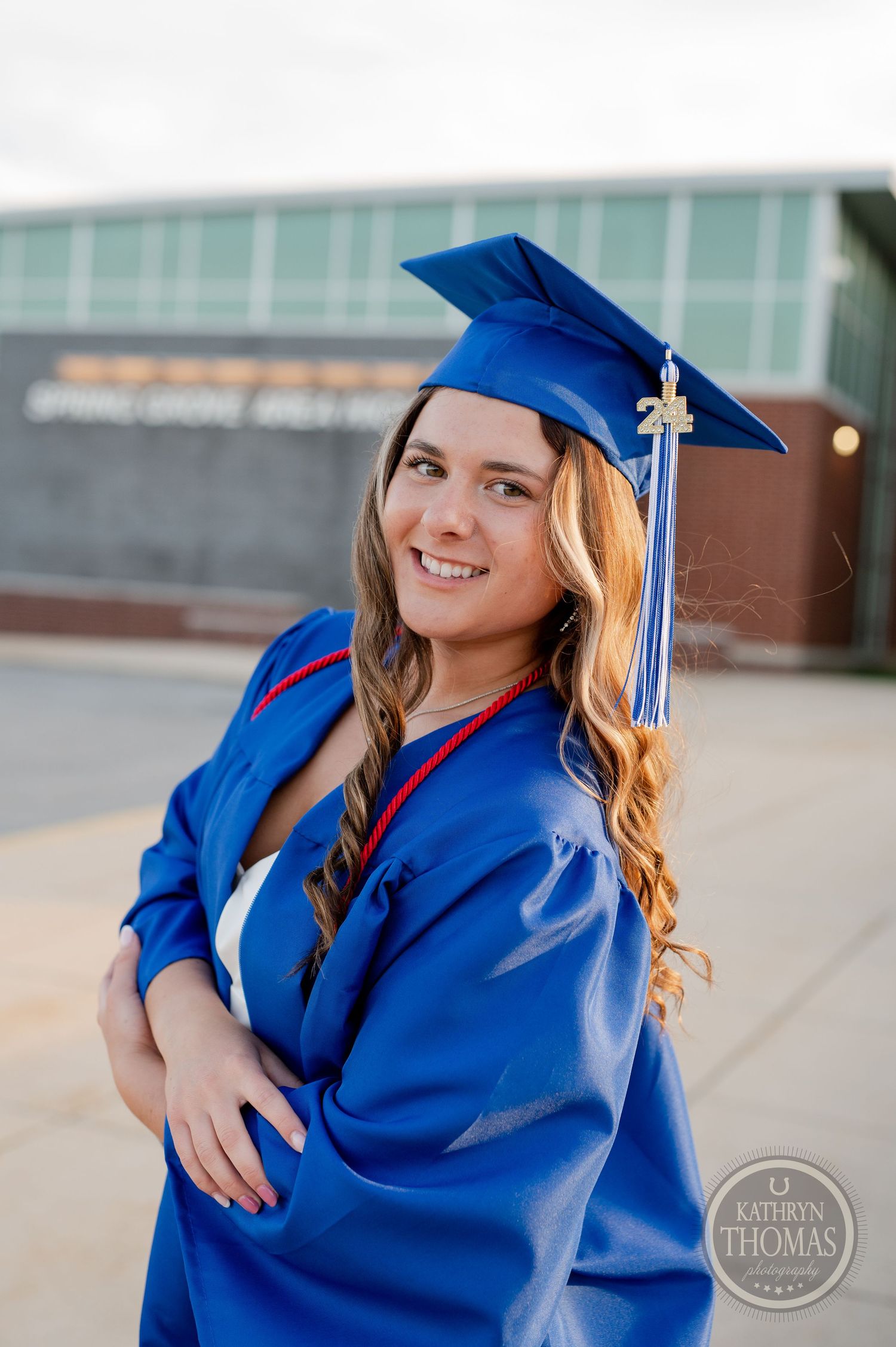 Graduate in blue cap and gown poses in front of modern school building.